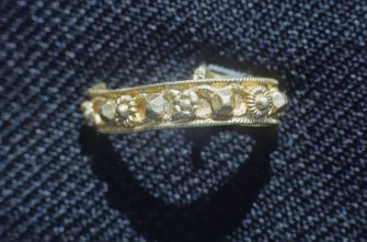Flat banded finger ring with spiralled beading at both edges. The central field is 	decorated with applied gold motifs of truncated prisms, pellet-bordered discs, six-	petalled rosettes, and daisy-like flowers. No mark present.