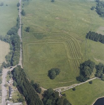 Oblique aerial view centred on the remains of the Roman Fort, chapel and burial ground, with Roman Temporary Camp and road bridge adjacent, taken from the SSW.