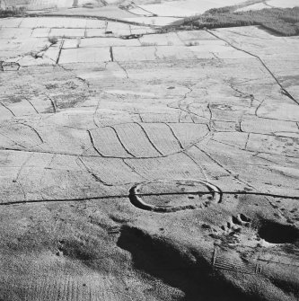Oblique aerial view of Kirk Hill centred on the remains of a fort and palisaded enclosure with buildings, enclosures, lazy-beds, quarries and a farmstead, field-system, rig and boundary bank adjacent, taken from the NW.