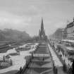 General view of Princes Street looking westwards showing the Castle, Mound, Scott Monument, Waverley Gardens and a busy street with trams, pedestrians and cars.