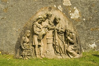 View of carved panel with baptism scene in churchyard to E of church