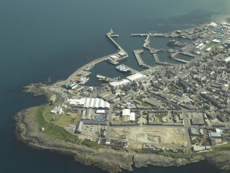 General oblique aerial view centred on the harbour with the lighthouse and remains of the tower in the foreground, taken from the NW.