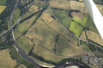 General oblique aerial view centred on the cropmarks of the Roman fort, Roman temporary camps and fort annexes with the railway viaduct and road bridge adjacent, taken from the NW.