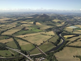 General oblique aerial view centred on the cropmarks of the Roman fort and Roman temporary camps with the town, village and remains of the fort in the distance and the railway viaduct and road bridge in the foreground, taken from the NE.