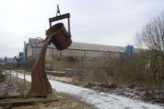 Voew of sculpture alongside public path of line of dismantled railway with back view of foundry buildings from south.