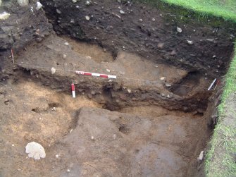 Trench 14 from the NW showing section excavation through the lower burnt mound deposit C13.27 and NW facing baulk of trench and features cut through the burnt mound deposit (scales = 0.5m and 0.2m)