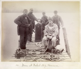 Diver in diver suit sitting on the pier with a group of men at Dalerb Slip, Kenmore', Loch Tay. Equipment and ropes to guide the diver can be seen.

