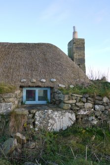 Detailed view of the Southern window to the West elevation of the thatched cottage at 13 Kilmoluaig, Tiree. The image shows the splayed window opening within the thick, battered wall. Stones tied to rope and 'chicken wire' can also be see above the window, which are for the purpose of keeping the thatching in place.