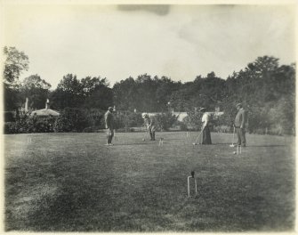 View of garden buildings and croquet lawn with group of people playing croquet, probably at Inchrye Abbey.

