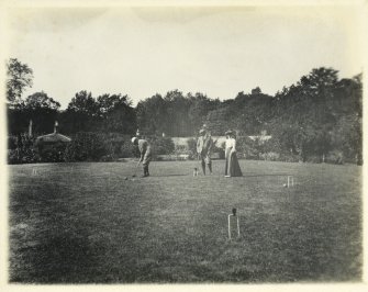 View of garden buildings and croquet lawn with group of people playing croquet, probably at Inchrye Abbey.
