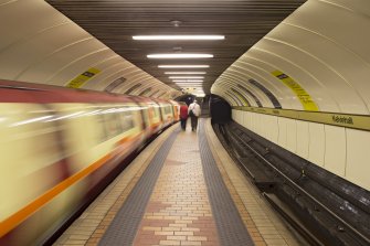 Interior. View looking along island platform and tracks of Kelvinhall subway station, with train leaving the station