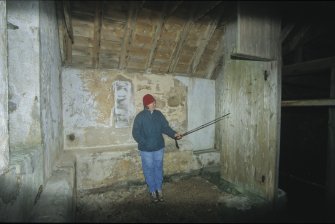 The small room in the south wing was probably a private facility for the estate gentry. The three outside walls were plastered, incorporating a plastered stone bench. The fourth wall consisted of tongued and grooved boarding. The figure is pointing to a boat graffito scratched on the wood. This wall has since been removed.