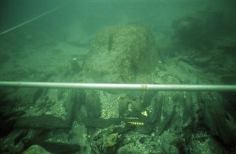 The displaced timbers surrounding this large rock are fragmented pieces of the hull, showing that the rock was in this position when the sinking vessel struck it, causing major damage to the port bow quarter. No timbers extend under the rock. Gun 6 is visible on the upper right. Metre graduations are visible on the grid-pole. (Colin Martin)