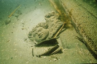 Carved wooden putto or cherub (DP92/169) exposed at the time of the Archaeological Diving Unit’s visit in 1992. In the foreground is a partly-exposed staved wooden costrel, and between it and the cherub is a human ulna. Wreck timbers are seen on the right. (Archaeological Diving Unit)
