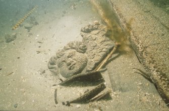 Carved wooden putto or cherub (DP92/169) exposed at the time of the Archaeological Diving Unit’s visit in 1992. In the foreground is a partly exposed staved wooden costrel (DP92/063) and between it and the cherub is a human ulna. Wreck timbers are seen on the right. (Archaeological Diving Unit)