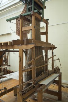 Interior. General view. Jaquard loom in gallery area. The warp threads in tension on the loom frame are clearly visible as is the jacquard device above the loom which manages the weft threads (by means of punched cards) to create the structure and pattern of the cloth being woven. The holes and blanks on the cards allow the hooks in the frame to select or not select threads to create the complex patterns in the cloth