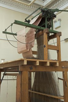 Interior. General view. Jaquard loom in gallery area. The warp threads in tension on the loom frame are clearly visible as is the jacquard device above the loom which manages the weft threads (by means of punched cards) to create the structure and pattern of the cloth being woven. The holes and blanks on the cards allow the hooks in the frame to select or not select threads to create the complex patterns in the cloth