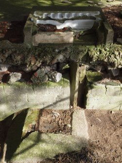 A urinal tray in the washroom of a barrack block with the remains of a timber post and a small drain in the foreground