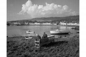 General view of Port Ellen across bay from South East, with couple sitting on bench in foreground.