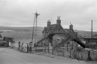 View from NNW showing footbridge with down platform building in background and level crossing gates in foreground