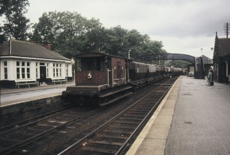 View from ESE showing goods train in station with down platform shelter on the left and footbridge in background