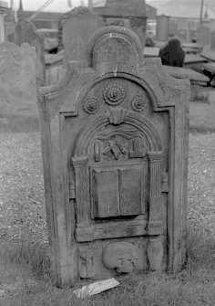 View of standing tombstone, St Mary's Church burial ground, Banff, with three rosettes at top, mason's symbols above opened book inserted in classical archway and hour-glass, skull and urn at bottom.