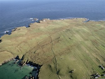 General oblique aerial view looking across the remains of the lazy beds and the turf-walled buildings towards the lighthouse, taken from the SW.