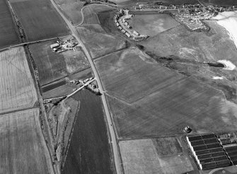 Oblique aerial view centred on the cropmarks of the ring ditch, pits and windmill at Sandend with Glenglassaugh Distillery adjacent, looking to the WNW.