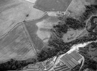 Oblique aerial view centred on the cropmarks of rig, a possible enclosure or long barrow at Rothes with Glenrothes Distillery adjacent, looking to the SW.

