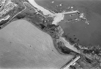 Oblique aerial view centred on the remains of the promontory fort at Castle Rock, Auchmithie with the harbour and coastguard station cottages adjacent, looking to the ENE.