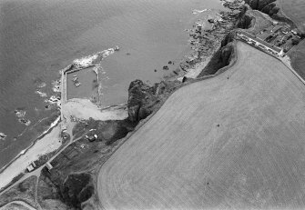 Oblique aerial view centred on the remains of the promontory fort at Castle Rock, Auchmithie with the harbour and coastguard station cottages adjacent, looking to the ESE.