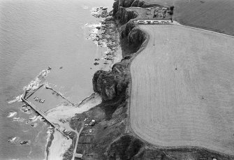 Oblique aerial view centred on the remains of the promontory fort at Castle Rock, Auchmithie with the harbour and coastguard station cottages adjacent, looking to the SE.