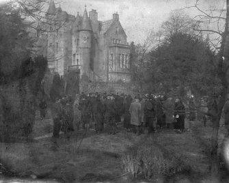 General distant view of the house partially obscured by trees with a large group of people walking around the grounds, seen from the South.