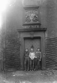 Detailed exterior view of the entrance doorway with three workmen standing in it seen from the West North West. Above the studded door is a stone relief of a coat-of-arms of two lions flanking a crest dated 1831 and with the initials TDL and CAC.