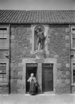 South elevation of central portion of Alexander Selkirk's former house, with woman standing in doorway and 1885 statue of the mariner from Fife above.