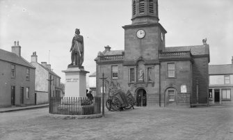 View from S of town hall and statue of Robert Bruce, High Street, Lochmaber. Large carts are balanced end on behind the statue.

