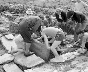 Veiw of the Middleton family, Annie Gillies and RCAHMS staff lifting a grave marker at Inchmarnock chapel in 1975, taken from the W.