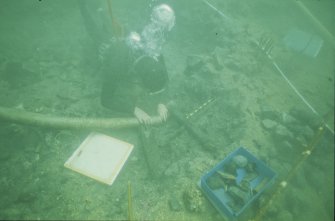 Paula Williams carrying out area excavation within the site grid. A wooden caulking mallet is being cleaned for recording - note her hand held over the nozzle for critical suction control. Drawing-board and finds box are to hand.