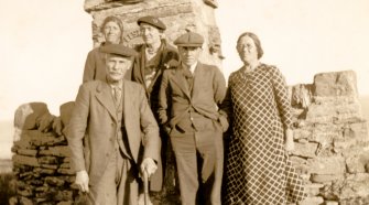 Scanned image of photograph of two men and three women standing in front of monument and stone wall at Nybster