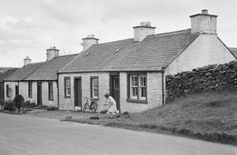 View of south side of Victoria Street, Kirkpatrick Durham, from north west, showing a woman cutting grass outside no.53