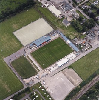 Oblique aerial view centred on the football stadium with courthouse, police station and hospital adjacent, taken from the NW.