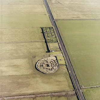 Oblique aerial view centred on the remains of the broch with the memorial and graveyard adjacent, taken from the SW.