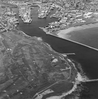 Oblique aerial view centred on the remains of the coastal battery with breakwaters adjacent and harbour in background, taken from the E.