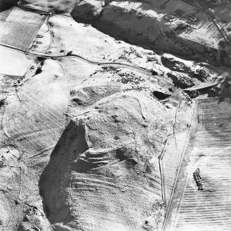 Oblique aerial view centred on the remains of the fort, hut-circles and cairn with hut-circle, enclosures, building and rig adjacent, taken from the NW.