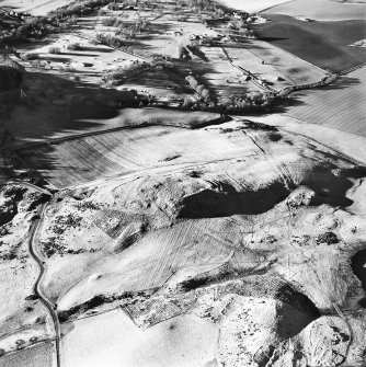 Oblique aerial view centred on the remains of the fort, hut-circles, cairn, building, rig and enclosures with enclosures adjacent, taken from the E.