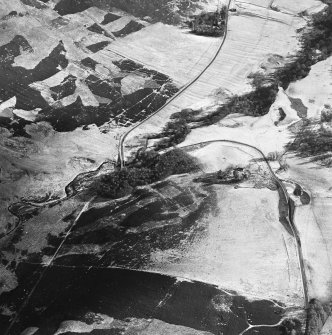 Oblique aerial view centred on the remains of buildings and enclosures with road bridge, rig and farmsteading adjacent, taken from the NW.