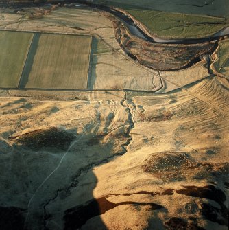 Oblique aerial view centred on the remains of the enclosure with the remains of the homestead, buildings, rig and cultivation terraces adjacent, taken from the SE.