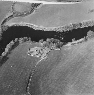 Oblique aerial view centred on the remains of the church and burial ground with the remains of a motte in trees adjacent, taken from the S.