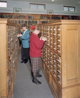 Interior.  View within library showing wooden cabinets containing drawers of index cards, cross-referencing Reports with reference books,journals and other records.  A large part of the library's holdings was sent to ICI Explosives headquarters in Australia in 1996, but the Index remained in Ardeer.  Also in the picture are Annette Kelly (right, Information Officer) and Sheila Conway (former Records and Reports Officer)