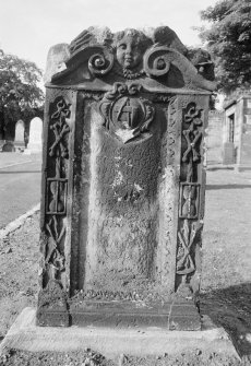 View of gravestone commemorating James Lawson 1728 in Calton Burial Ground, Edinburgh. The stone has a scrolled broken pediment with a central putto face with two skulls on the corners of the stone, and border of ribbands tied around crossed bones, an hourglass, scythe and pick, (the emblems of death), and a central cartouche holds the emblems of a mason.